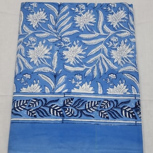 May include: Folded blue fabric with a white floral pattern and a dark blue leaf border. The fabric has a solid blue border at the bottom. The floral design includes large white flowers and white leaves.