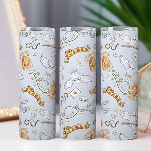Puede incluir: Tres vasos de color azul claro con un patrón repetido de búhos, bufandas, gafas y escudos. El diseño presenta detalles dorados, rojos y amarillos. Los vasos son cilíndricos y parecen estar hechos de metal o un material similar.