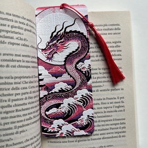 Può includere: Segnalibro decorativo con un drago rosso e viola, davanti a una luna piena e onde stilizzate. Il drago è ornato da un fiocco rosso. Ideale per gli amanti della lettura.