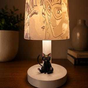 May include: A white table lamp with a cylindrical base and a lampshade featuring a black and white anime illustration. A black cat figurine with a crescent moon on its forehead sits on the base. The lamp is on a wooden surface.
