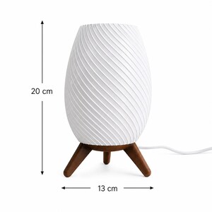 Può includere: Una lampada da tavolo bianca a forma di uovo con un motivo a strisce diagonali strutturate. La lampada è alta 20 cm e larga 13 cm, sostenuta da tre gambe in legno marrone. Un cavo di alimentazione bianco si estende dalla base.