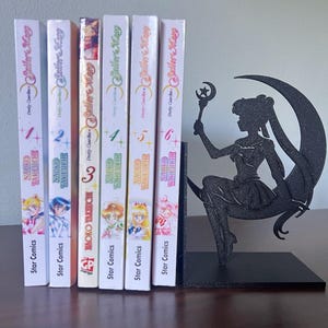 Può includere: Una collezione di sei volumi del manga Sailor Moon, con una silhouette nera decorativa di un personaggio di Sailor Moon che tiene una bacchetta, usata come fermalibri. I dorsi dei manga sono bianchi con illustrazioni e testo colorati.