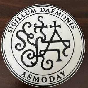 Puede incluir: Un emblema redondo, blanco y negro, con el texto "SIGILLUM DAEMONIS" alrededor del borde. El centro presenta un símbolo negro complejo con la palabra "ASMODAY" en la parte inferior. El diseño es una ilustración gráfica detallada.