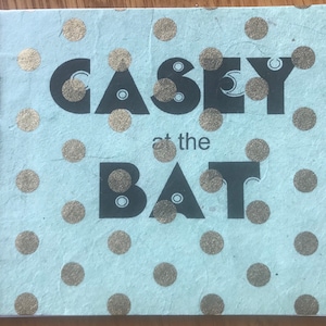 Puede incluir: Un libro azul claro con lunares dorados y el texto "Casey at the Bat" en letras negras.