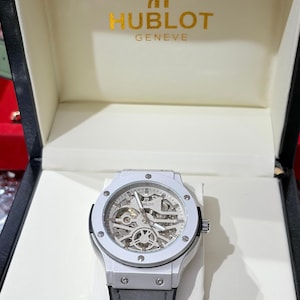 Op de afbeelding: Een witte Hublot Geneve horloge met een grijze leren band, gepresenteerd in een merkdoos. Het horloge heeft een skeleton wijzerplaat, die de interne mechanismen onthult. De horlogekast is wit en de band is grijs.