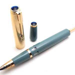 May include: A vintage fountain pen with a gold-colored cap and accents, a teal barrel, and a gold nib. The cap is off, revealing the nib and the teal barrel. The pen has a blue jewel on the cap and the top of the barrel.