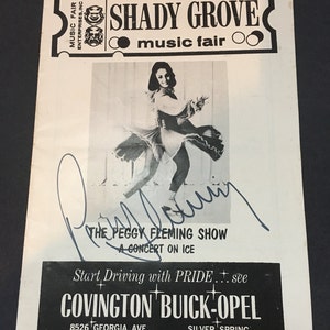May include: Black and white photo of a woman ice skating in a white dress. The photo is signed "Peggy Fleming" and is advertising "The Peggy Fleming Show - A Concert on Ice" at the Shady Grove Music Fair. The ad also includes information for Covington Buick-Opel, located at 8526 Georgia Ave, Silver Spring, 587-7300.