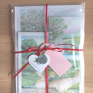 Può includere: Un set di biglietti d'arte paesaggistica, avvolti in cellophane trasparente e legati con un nastro rosso. I biglietti presentano dipinti ad acquerello di alberi, campi e un sentiero. Un'etichetta a forma di cuore con il testo "angelimcart.com" è allegata.
