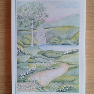 Può includere: Quaderno con un dipinto ad acquerello di un paesaggio sereno. L'opera raffigura un sentiero tortuoso, un piccolo stagno e dolci colline verdi sotto un cielo rosa e blu tenue. La parola "Notebook" è stampata in basso.
