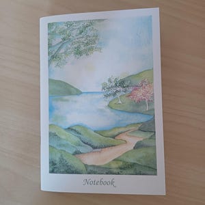 Può includere: Un taccuino con una scena di paesaggio ad acquerello. L'immagine presenta un lago sereno circondato da colline e alberi verdi, con un sentiero che conduce all'acqua. La parola "Notebook" è stampata in basso.