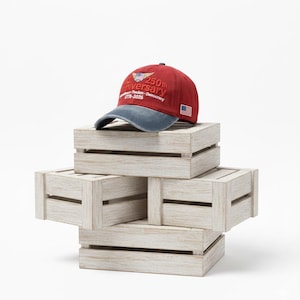 May include: A red and blue baseball cap with the text "250th Anniversary" and an American flag patch. The hat is displayed on a stack of four white wooden crates.