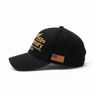 May include: Black baseball cap with a curved brim. Features gold embroidered text that reads "250th Anniversary Freedom Democracy 2026" and a small American flag patch. The cap is a classic style.