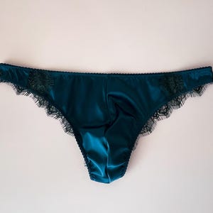 Op de afbeelding: Petrolkleurige satijnen slip met delicate zwarte kanten rand. Het lingerie heeft een laag uitgesneden model en een gladde, glanzende textuur. De kanten details geven het kledingstuk een vleugje elegantie.