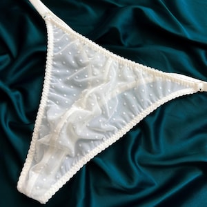 Op de afbeelding: Een witte string gemaakt van doorschijnende mesh-stof met een delicaat stippenpatroon. Het kledingstuk is afgewerkt met een geschulpte rand langs de tailleband en beenopeningen, en bevat verstelbare bandjes met kleine witte metalen onderdelen.