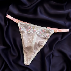 Könnte beinhalten: Ein Tanga mit beiger Basis und weißen Punkten, akzentuiert durch ein oberes Paneel aus transparenter weißer Spitze, das mit kleinen rosa Rosen und grünen Blättern bestickt ist. Das Kleidungsstück hat verstellbare hellrosa Elastikbänder.