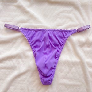 Peut inclure: Un string violet vif avec une texture côtelée, des bretelles latérales fines et réglables dotées d'anneaux métalliques argentés, et une poche avant profilée. Le vêtement présente des coutures délicates le long des bords du tissu.