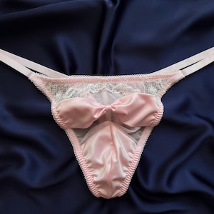 May include: A light pink thong featuring a satin finish, a central satin bow, and white lace trim along the top edge. The garment includes thin, adjustable side straps and a sheer mesh panel, constructed with delicate scalloped edges throughout.