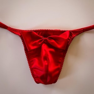 May include: A vibrant red thong made of satin fabric. The thong features a large bow at the front and delicate trim along the edges. The smooth, shiny material reflects light, highlighting the details of the garment.