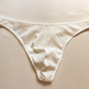 May include: White thong underwear with a delicate, textured fabric. The edges are trimmed with a scalloped lace detail. The fabric has a subtle striped pattern.
