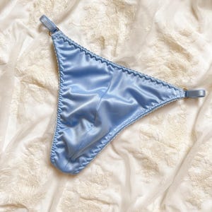 Könnte beinhalten: Hellblauer Satin-String mit gewelltem Rand. Die Dessous haben dünne Träger und ein klassisches String-Design. Der Stoff hat einen dezenten Glanz, und das Kleidungsstück wird auf weißem Stoff mit floralem Muster präsentiert.