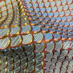 May include: Close-up of iridescent, scale-like material in shades of gold, green, and blue. The scales have a textured, striped pattern and are arranged in a layered, overlapping design. The background is a warm, reddish-brown color.