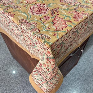 May include: A rectangular tablecloth featuring a floral design in gold, red, and green hues. The tablecloth has a detailed border and is draped over a wooden surface. The corner of the cloth is visible, displaying the intricate pattern.