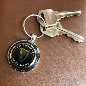 May include: A silver key chain with a black and gold round bottle opener. The bottle opener has the Guinness harp logo and the text "Use opener to enjoy safely".