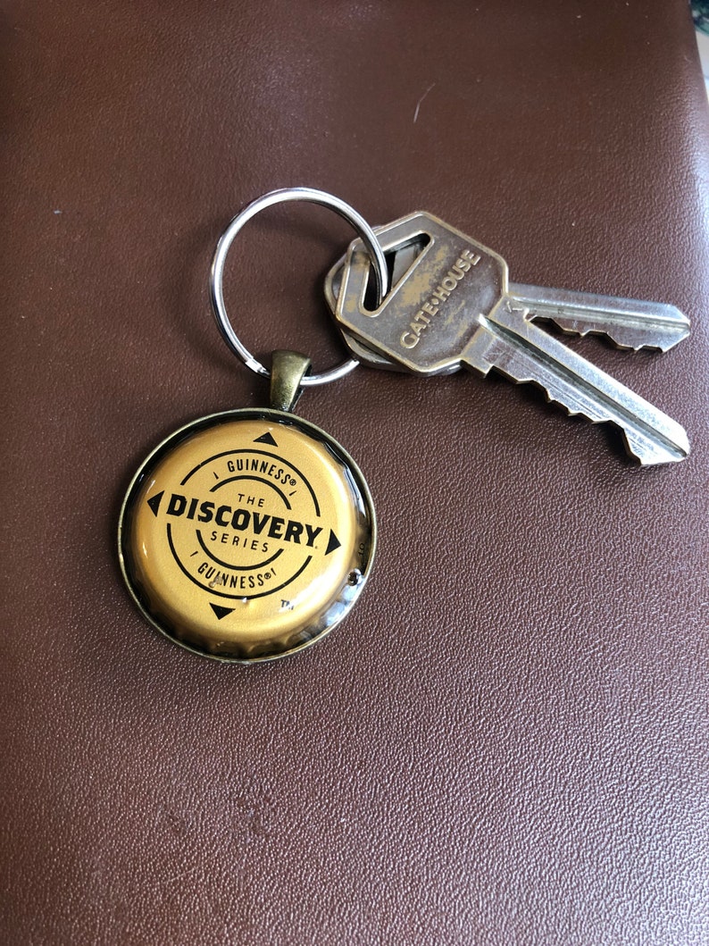 May include: A gold-colored key chain with a round, gold-colored bottle cap charm. The charm has the words "Guinness The Discovery Series" printed on it. The key chain also has two silver keys with a gold-colored key tag that says "Gate House".