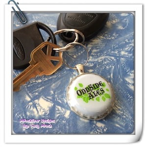May include: A keychain with a bottle cap charm featuring the text "Oddside Ales" in black lettering with green leaves on a white background.