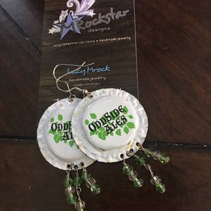 May include: A pair of earrings made from upcycled bottle caps with green glass beads hanging from each. The bottle caps are white with green text that reads "Oddside Ales".