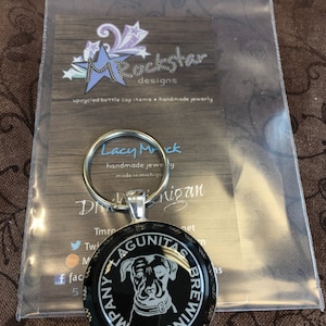 May include: A silver key chain with a black and white bottle cap charm featuring the Lagunitas Brewing Company logo. The logo is a black and white illustration of a dog with the words "Lagunitas Brewing Company" around the outside.
