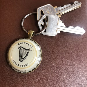 May include: A gold-colored keychain with a white Guinness Extra Stout bottle cap charm. The bottle cap features the Guinness harp logo and the text "GUINNESS EXTRA STOUT Est. 1759 TM".