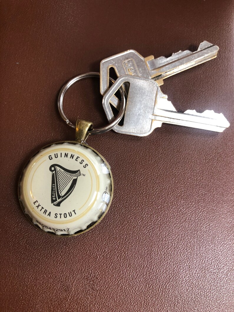 May include: A gold-colored keychain with a white Guinness Extra Stout bottle cap charm. The bottle cap features the Guinness harp logo and the text "GUINNESS EXTRA STOUT Est. 1759 TM".