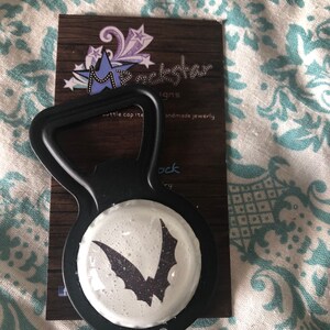 May include: Black bottle opener with a white circle featuring a black bat silhouette. The bottle opener is on a brown card with the text "M. Rockstar Designs" and "Handmade Jewelry".