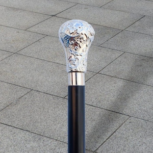 May include: A black walking stick with a silver-coloured, ornate handle. The handle features a detailed floral design. The stick has a silver-coloured band where the handle meets the black shaft. The stick is standing on a grey tiled surface.