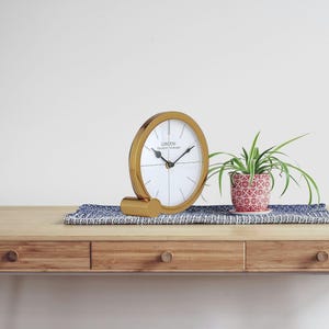 May include: A gold-coloured desk clock with a white face and black hands. The clock has the word "LONDON" printed on the face. A small potted plant with green leaves sits next to the clock on a blue patterned runner. The clock is on a wooden desk.