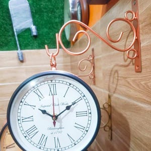 May include: A double-sided clock with a copper-colored metal frame and bracket. The clock faces are white with black Roman numerals and hands. The text "Imperial Clock Works" and "Birmingham at 1792" are printed on the clock face. The clock is mounted on a wooden wall.