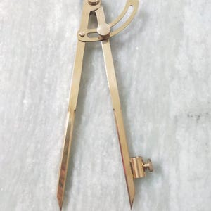 Könnte beinhalten: Ein messingfarbener Zirkel im Vintage-Stil mit spitzen Spitzen. Das Werkzeug hat einen verstellbaren Bogen und eine Schraube für präzise Einstellungen. Der Zirkel dient zum Messen von Entfernungen und zum Zeichnen von Kreisen, üblich beim Zeichnen und in der Geometrie.