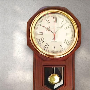 Könnte beinhalten: Eine Wanduhr aus Holz mit goldfarbenem Rahmen und römischen Ziffern. Das Zifferblatt ist cremefarben mit dem Text "OXFORD" und "Franklin & Marshall EST 1868". Unterhalb hängt ein goldfarbenes Pendel.