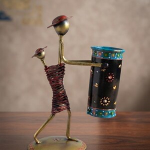 May include: A metal desk organizer featuring two stylized figures with wire-wrapped bodies and hats. The taller figure holds a black cylindrical container decorated with blue bands and small floral patterns. The entire structure stands on a circular base.