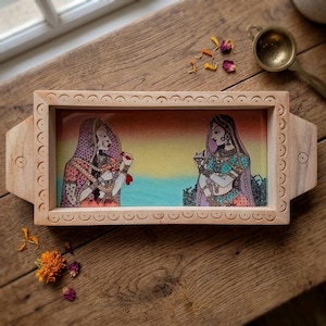 Puede incluir: Una bandeja de madera rectangular con asas, que presenta una ilustración pintada de dos figuras con vestimenta tradicional frente a frente sobre un fondo degradado en naranja, amarillo y azul, enmarcada por un patrón circular tallado.