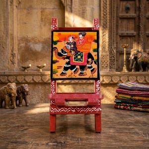May include: A red wooden book stand featuring a hand-painted square panel depicting two figures riding an elephant. The stand has white decorative patterns along its edges and a central horizontal support bar for holding books or tablets.