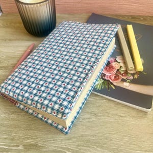 Könnte beinhalten: Ein Hardcover-Buch mit blau-weißem Karomuster und roten Herzakzenten. Ein rosa Stift, eine Kerze in einem blauen Glas und ein Notizbuch mit floralem Design sind ebenfalls zu sehen. Die Gegenstände sind auf einer Holzoberfläche angeordnet.