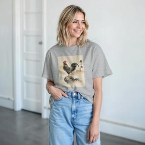 Op de afbeelding: Een lichtgrijs t-shirt met een hanenillustratie. Het kunstwerk toont een haan die op een rots zit met een berglandschap. De haan heeft rode, zwarte en grijze veren. Japanse tekst staat aan de rechterkant van de afbeelding.