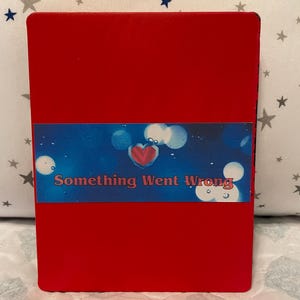 Puede incluir: Un objeto rectangular rojo con una banda azul y blanca con el texto "Something Went Wrong" y un corazón. El objeto está sobre un fondo blanco con un patrón de estrellas.