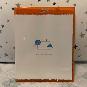 Op de afbeelding: Een oranje Blu-ray disc case met een witte papieren inleg. De inleg heeft een geometrisch ontwerp met de tekst "Something went wrong". De case ligt op een oppervlak met sterrenpatroon.