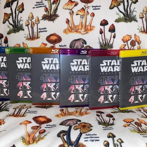 Puede incluir: Seis estuches Blu-ray Star Wars Project 4K con lomos de diferentes colores. Los estuches están dispuestos en fila, con el texto "STAR WARS" y "4K" visibles en las portadas. El fondo es una tela blanca con un estampado de setas.