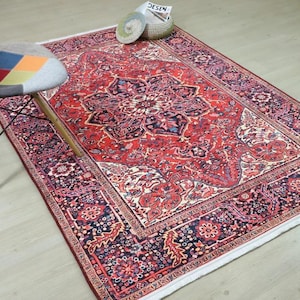May include: A rectangular, red Persian rug with an intricate floral pattern and a dark border. A multi-colored patchwork chair with wooden legs sits to the left of the rug. A woven basket and magazine are on the rug.