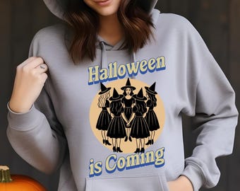 May include: A light gray hooded sweatshirt with the text "Halloween is Coming" and a graphic of witches. An orange pumpkin with a carved face sits to the left. The sweatshirt has a front pocket and is worn by a person.