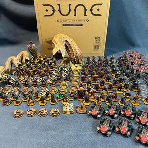 Könnte beinhalten: Eine Dune War for Arrakis Exclusive Edition Brettspielschachtel mit einer großen Sammlung bemalter Miniaturfiguren, darunter mehrere sandfarbene Sandwürmer, zahlreiche Infanterieeinheiten in verschiedenen Posen und schwarz-rote Panzerfahrzeuge.